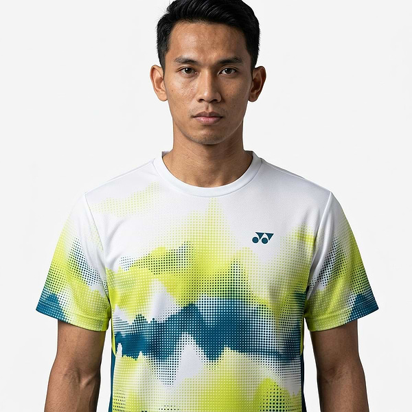 A young man with short black hair wears a Yonex YTM2025A Mens Badminton T-Shirt - White by Yonex, featuring green, blue, and yellow abstract patterns, standing against a plain white background and looking directly at the camera with a neutral expression.