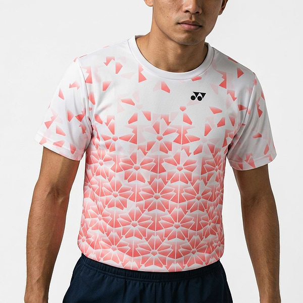 A person wears the Yonex YTM2025B Men’s Badminton T-Shirt in white, featuring a pink geometric floral pattern and small black logo, paired with dark shorts against a plain background.