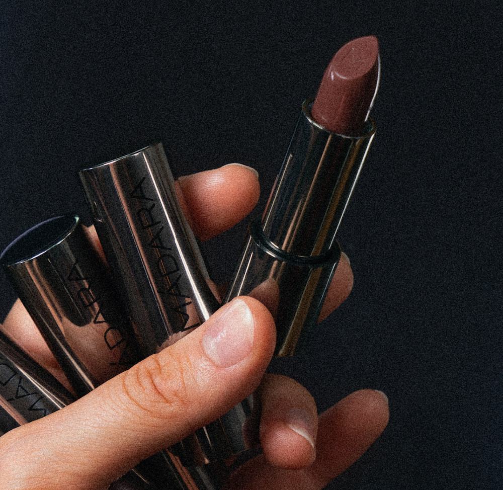 Close-up of a hand holding MÁDARA Dazzle Nights satin shine lipstick in a metallic tube, revealing a shimmering warm-toned shade