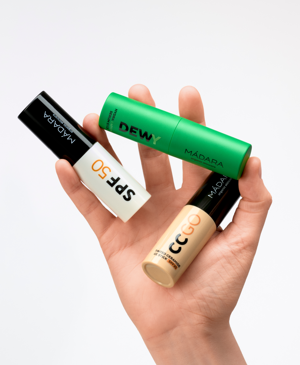 Hand holding three MÁDARA skincare sticks—DEWY Hydra Caffeine Serum Stick, SPF50 sunscreen stick, and CC GO tinted stick—against a clean light background