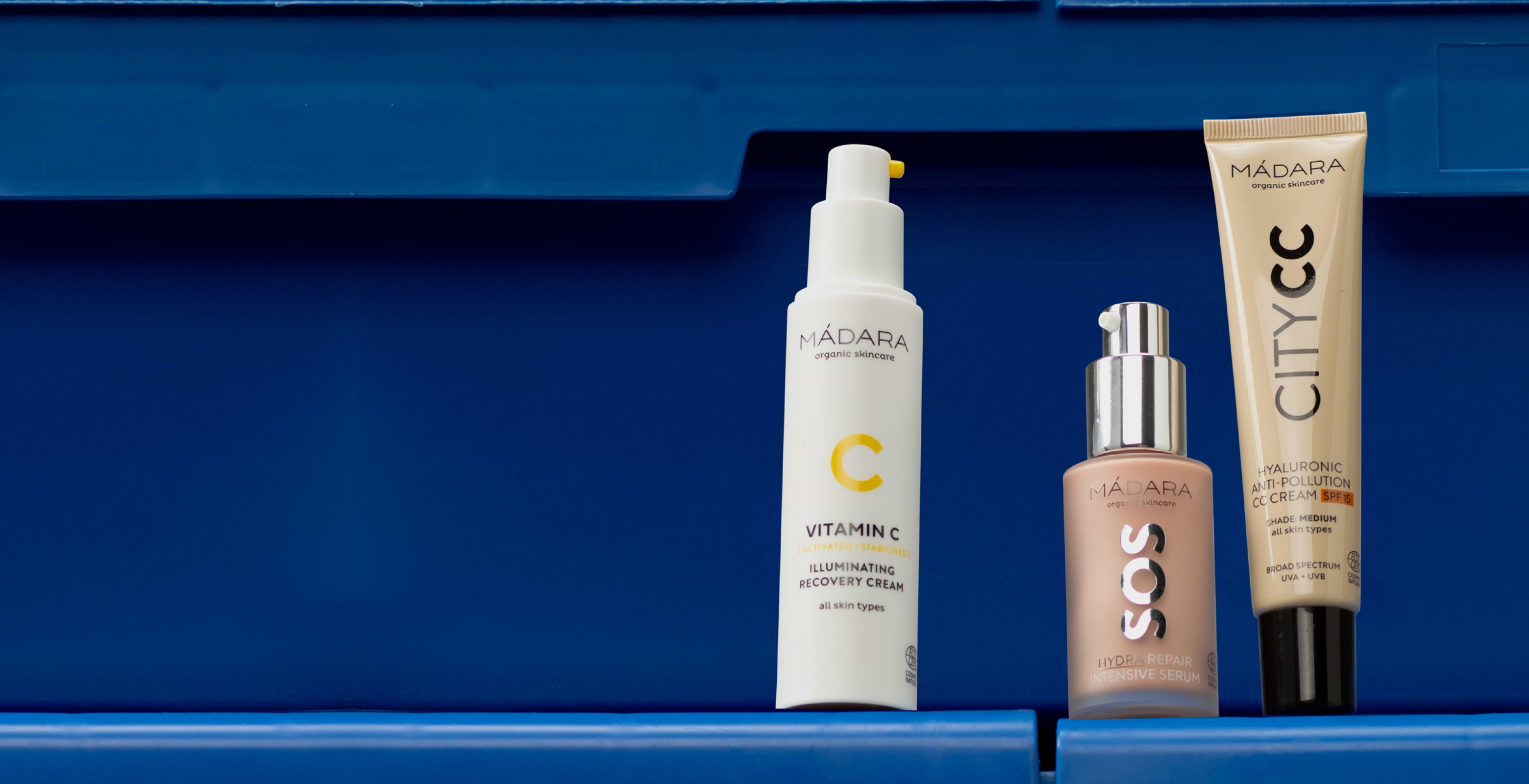 MÁDARA skincare products including Vitamin C Illuminating Recovery Cream, SOS Hydra Repair Intensive Serum, and City CC cream displayed against a bold blue background