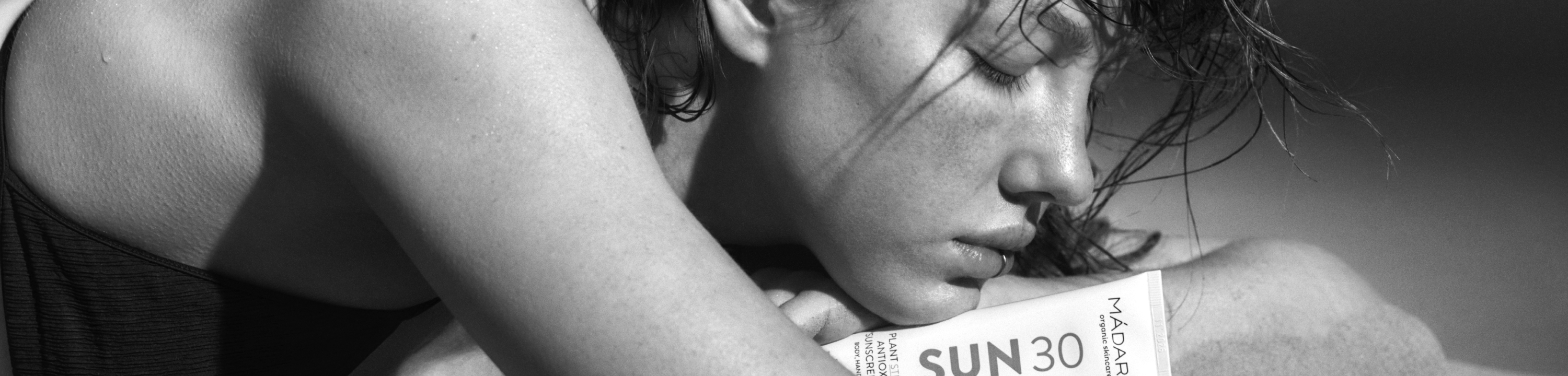 Black-and-white close-up of a person with closed eyes resting on their arms while holding a madara mineral sunscreen SUN30