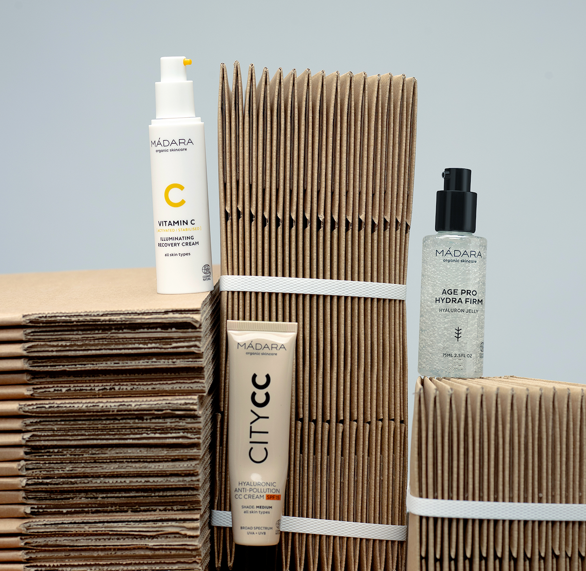MÁDARA skincare products including Vitamin C recovery cream, City CC cream, and Age Pro Hydra Firm hyaluron jelly displayed among stacked recycled cardboard packaging against a soft light background.