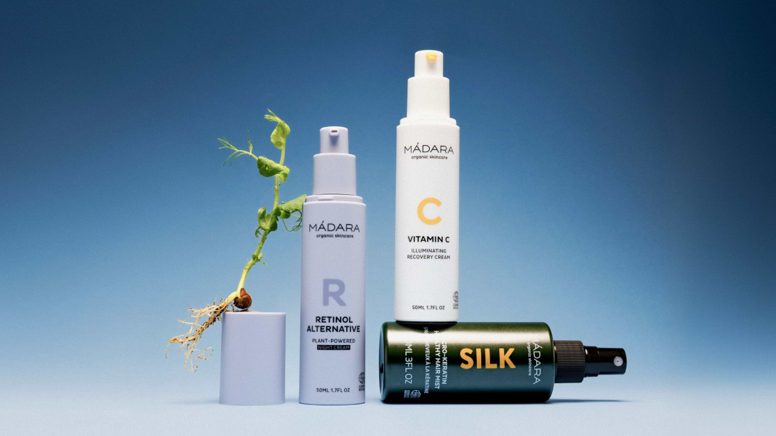 MÁDARA skincare products displayed on a blue background with a retinol alternative serum, Vitamin C cream, a silk hair product, and a small plant growing from a container