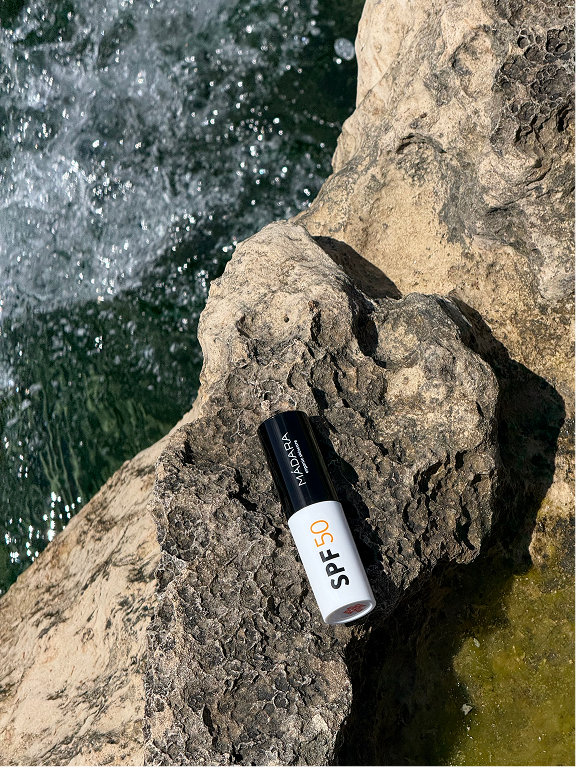 Madara SPF 50 sunscreen stick placed on a sunlit rock beside flowing water