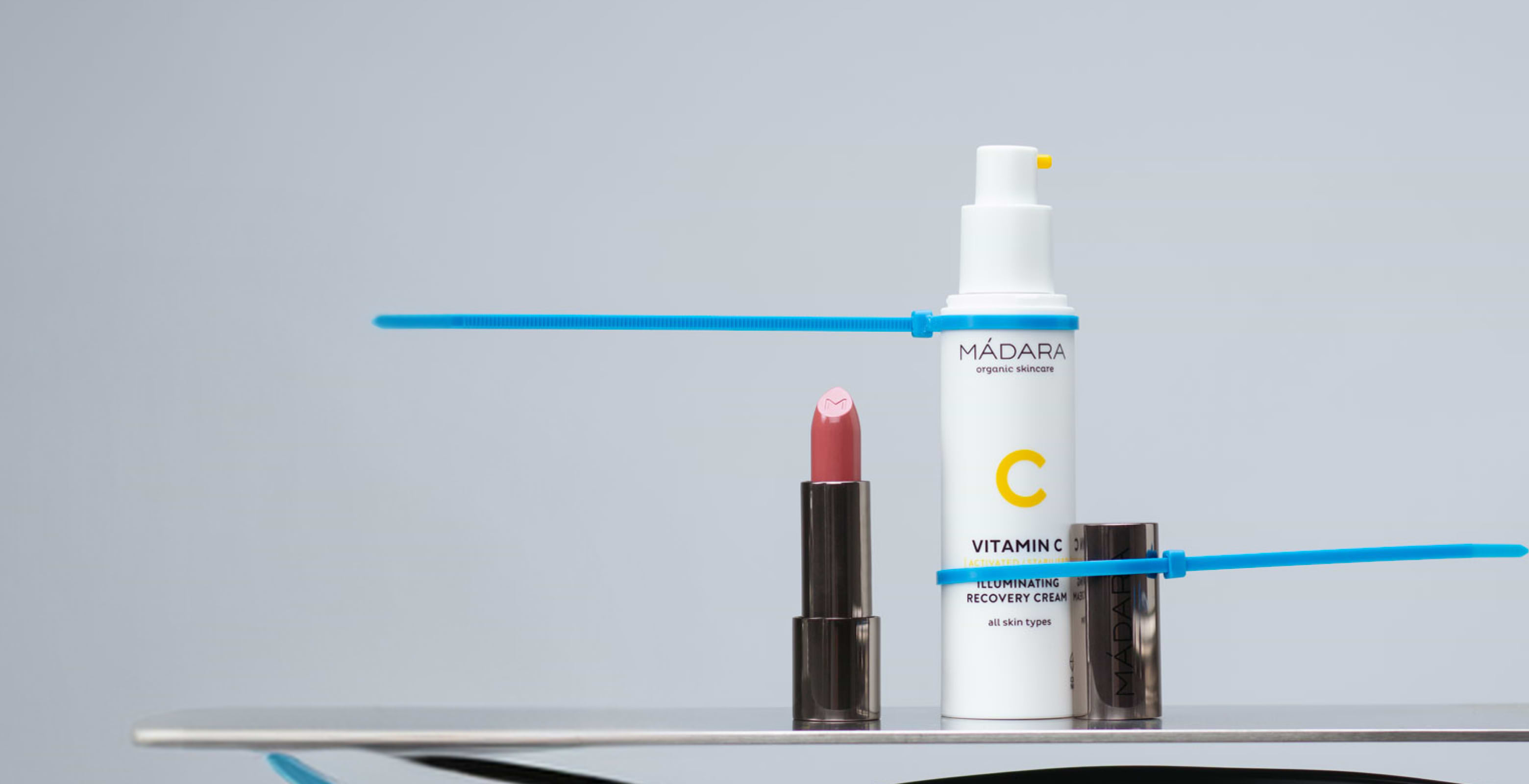 Minimal MÁDARA Vitamin C skincare and lipstick composition on a clean grey background with graphic blue accent