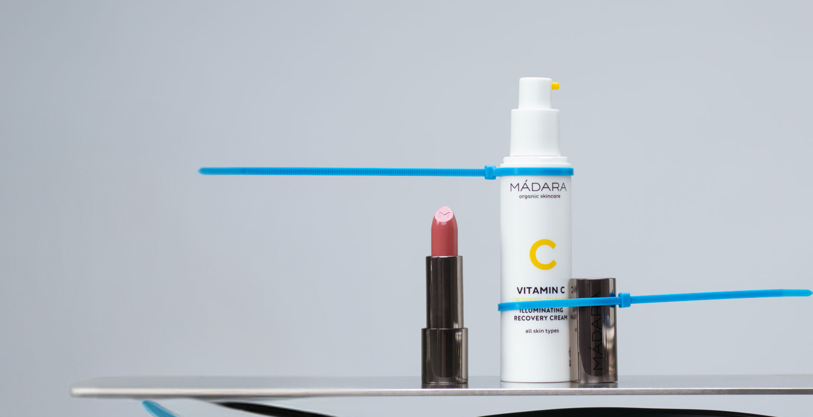 Minimal MÁDARA Vitamin C skincare and lipstick composition on a clean grey background with graphic blue accent