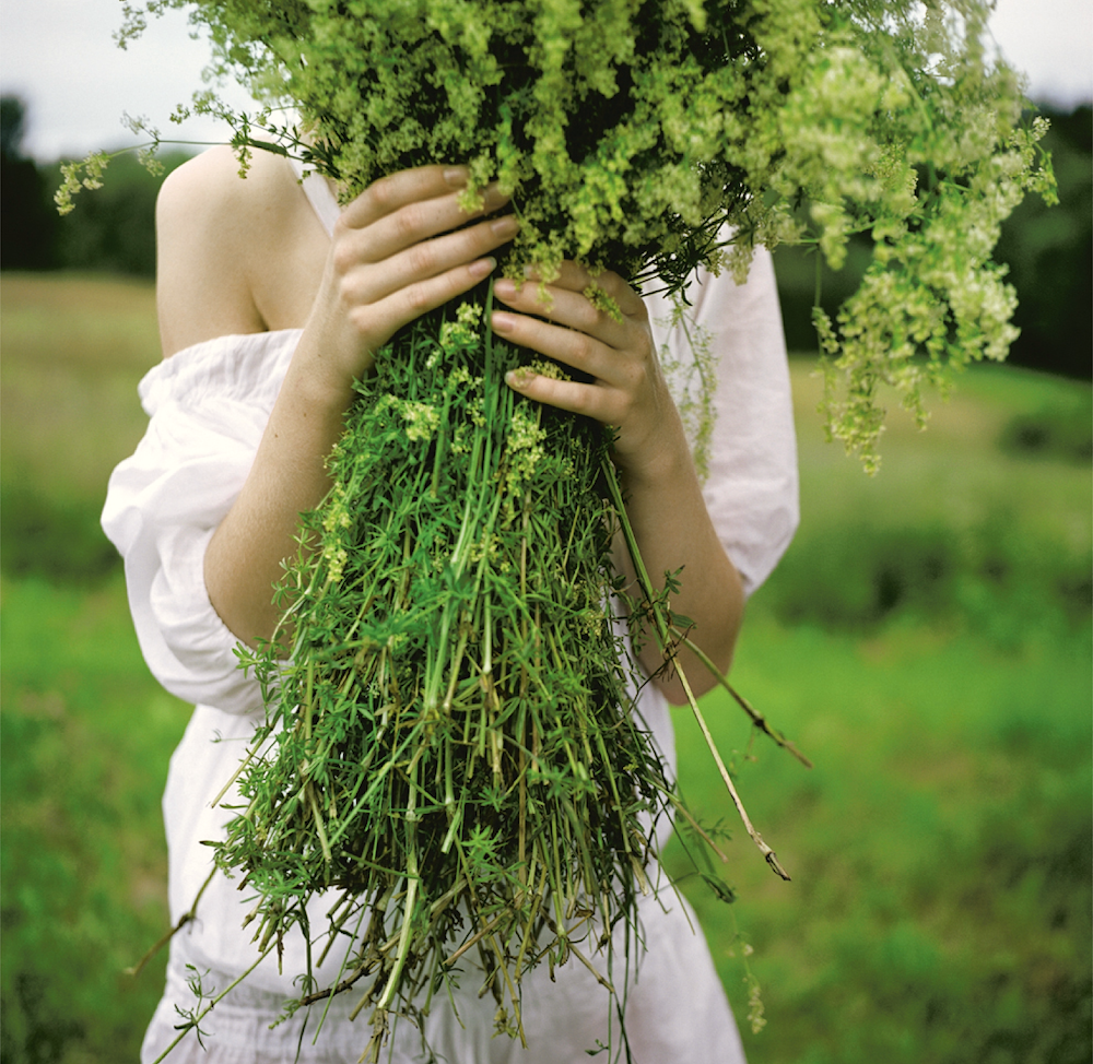 Person in a white dress holding a large bouquet of fresh green wildflowers, covering their face, in a grassy outdoor setting