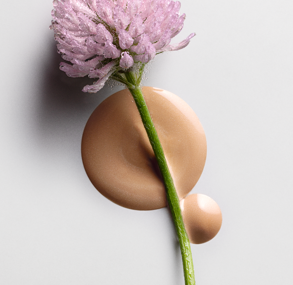Close-up of a dew-covered pink clover flower placed over smooth drops of beige tinted serum on a light background, representing the texture of MÁDARA Cosmetics H2O Hyaluron Tinted Serum in Sun Flower shade