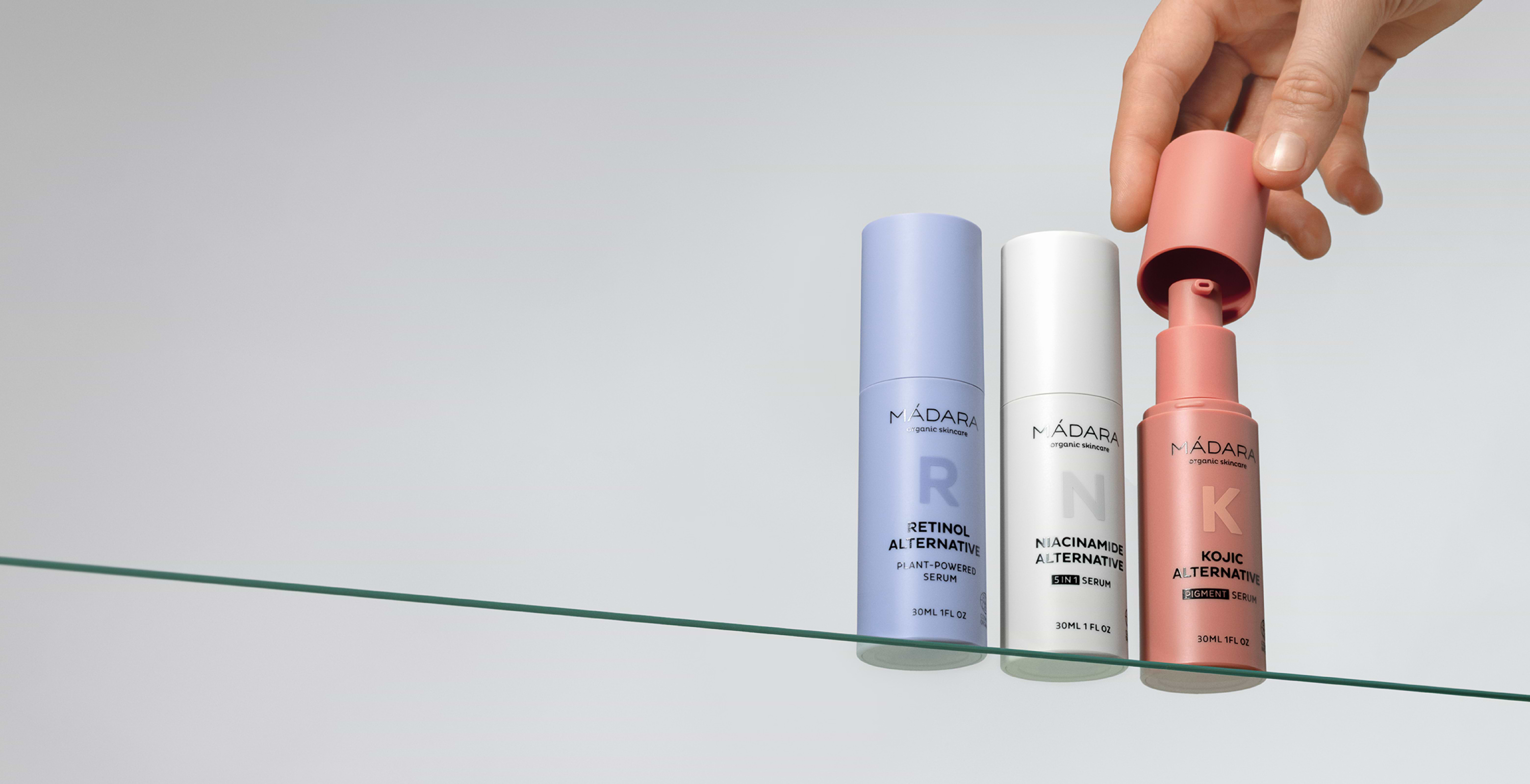 A hand removes the cap from a coral-colored serum bottle beside two other pastel bottles on a glass shelf, showcasing three facial serums from MÁDARA Organic Skincare: Retinol Alternative, Niacinamide Alternative, and Kojic Alternative, against a minimal light background