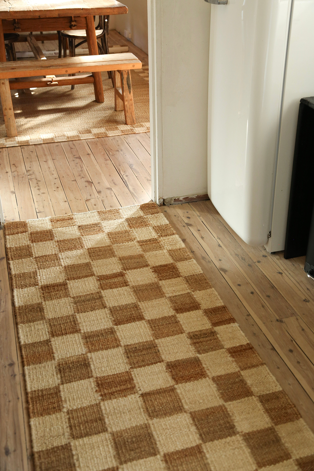 Checkered jute rug on a wooden floor with a wooden bench and white wall in the background.