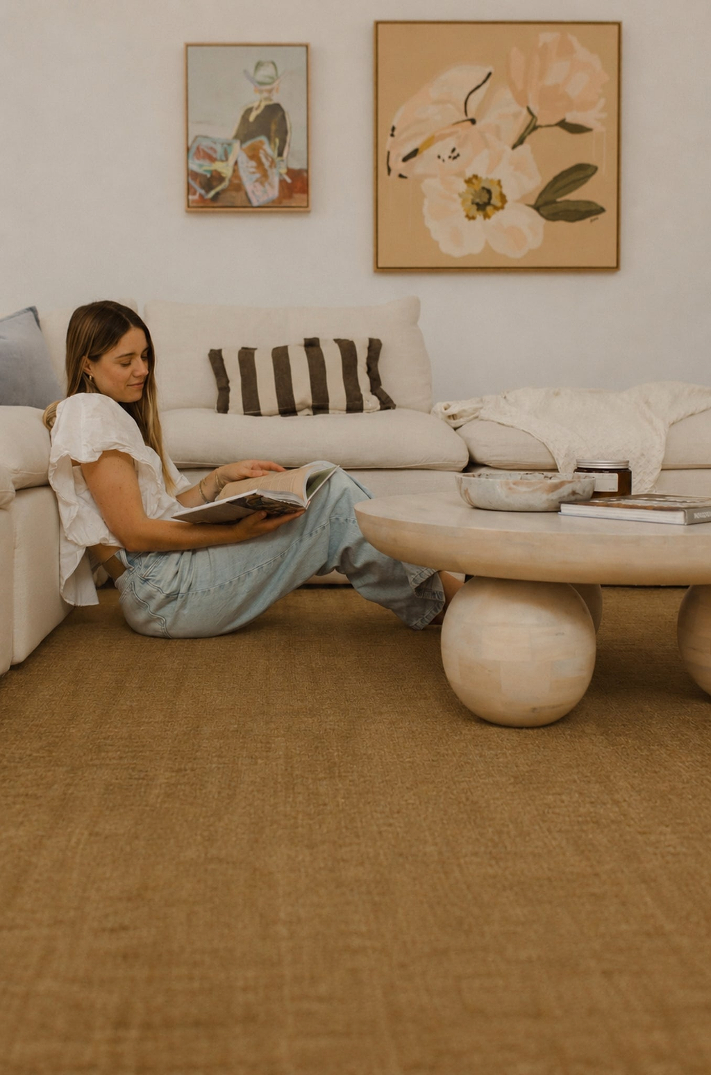 A woman sits on a cream sectional sofa in a living room, reading a book. The room features a prominent warm brown butter area rug by Rugs for Good, a round stone-topped coffee table, and two framed floral paintings on the wall.