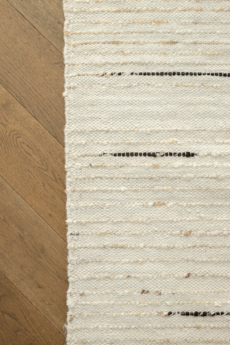 A close-up of a woven area rug with a textured cream and off-white striped pattern, featuring small black woven accents, shown next to a light brown wooden floor.