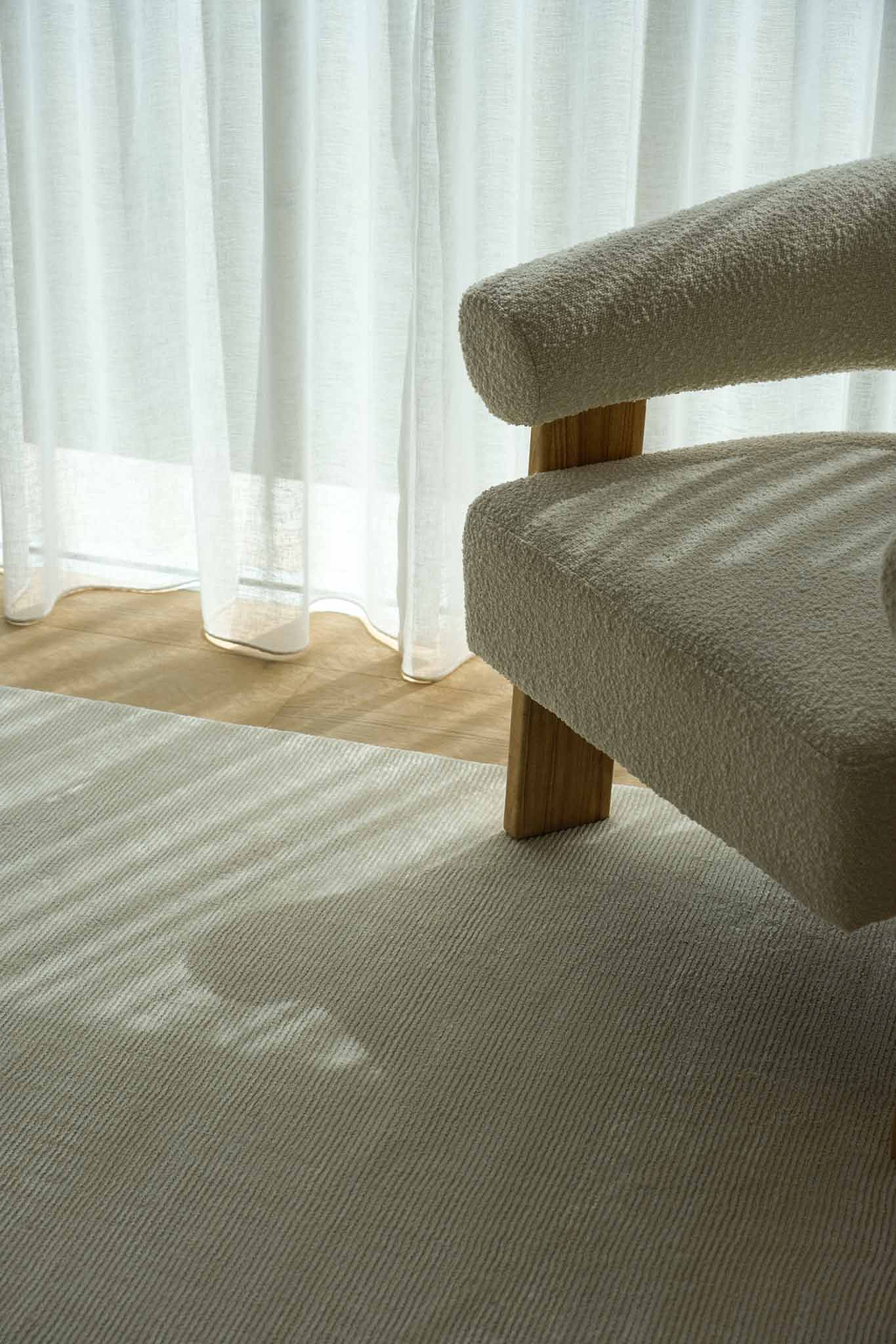 A close-up of a textured cream bouclé armchair with light wood legs, positioned next to sheer white curtains with soft window light casting shadows across the chair and a matching Yarra Linea cream rug by Rugs for Good