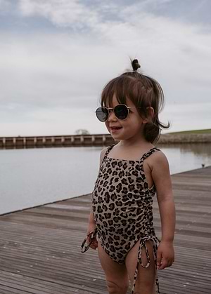 Child wearing Little Trouble leopard print one piece swimsuit and sunglasses on a wooden dock by a body of water.