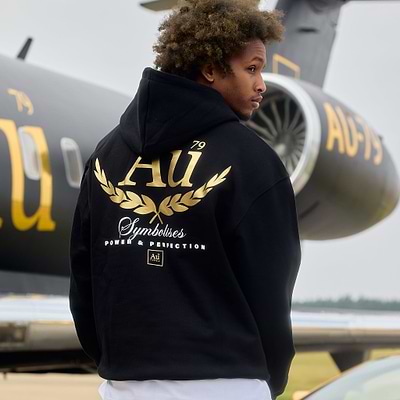 Person wearing a black hoodie with gold and white text and graphics, standing in front of a jet.