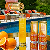 Hand pouring Au Vodka Tango Orange into a tall glass with two cans and oranges on a reflective surface