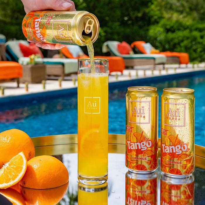 Hand pouring Au Vodka Tango Orange into a tall glass with two cans and oranges on a reflective surface