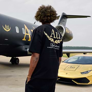 Person wearing a black t-shirt with gold lettering in front of a black airplane and gold car.