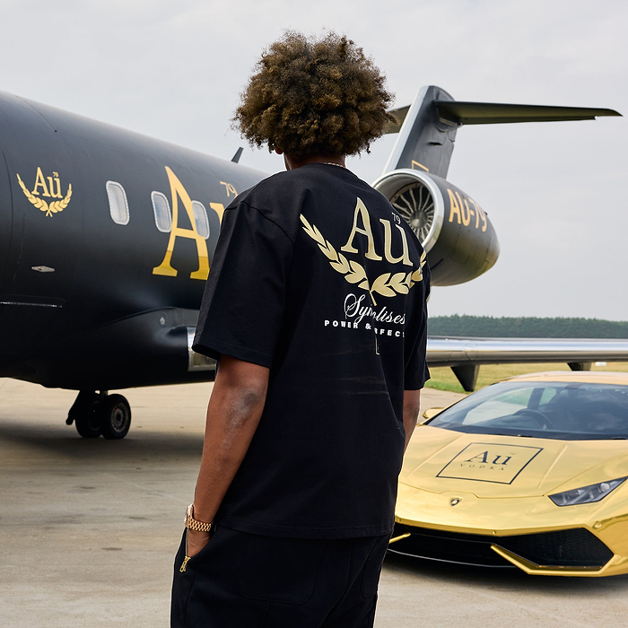 Person wearing a black t-shirt with gold lettering in front of a black airplane and gold car.