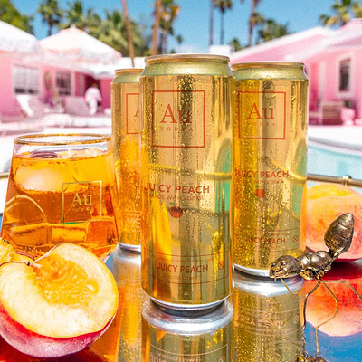 Cans of Au Peach sparkling water with a glass of the drink and peaches on a reflective surface.