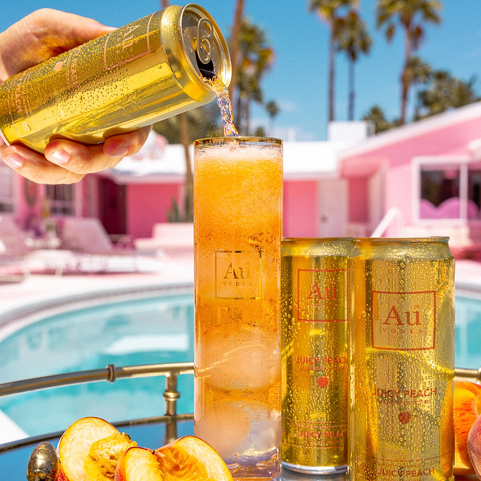 Person pouring a can of Au juicy peach into a glass with palm trees and pool in the background