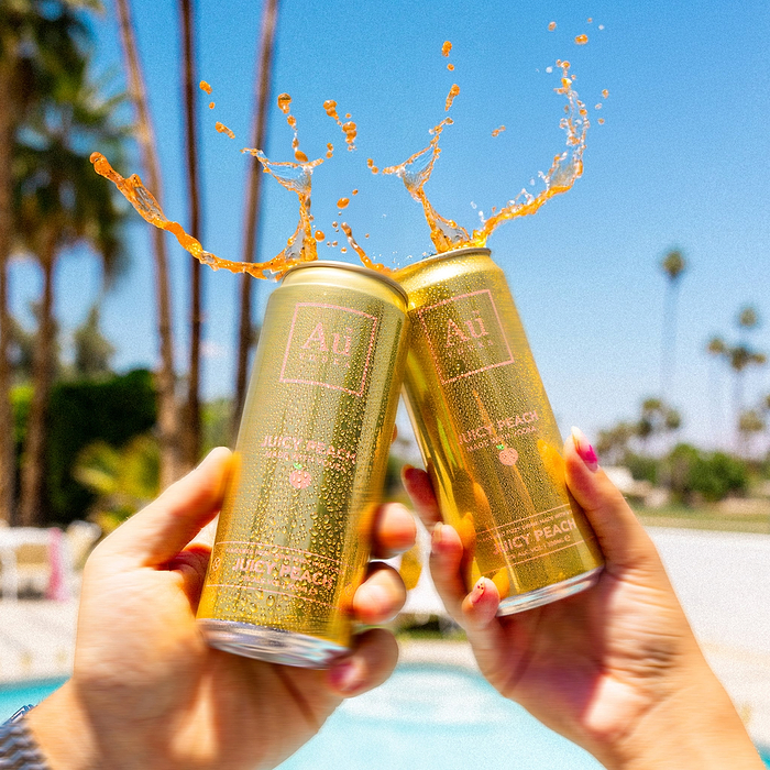 Two cans of Au Vodka juicy peach being clinked together with a splash against a poolside background.