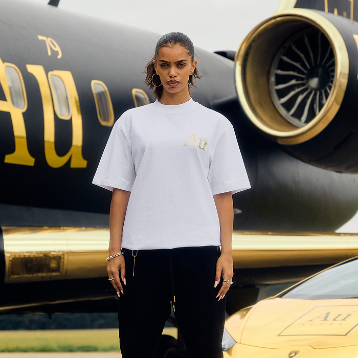 Person wearing a white t-shirt with gold logo in front of a black and gold airplane