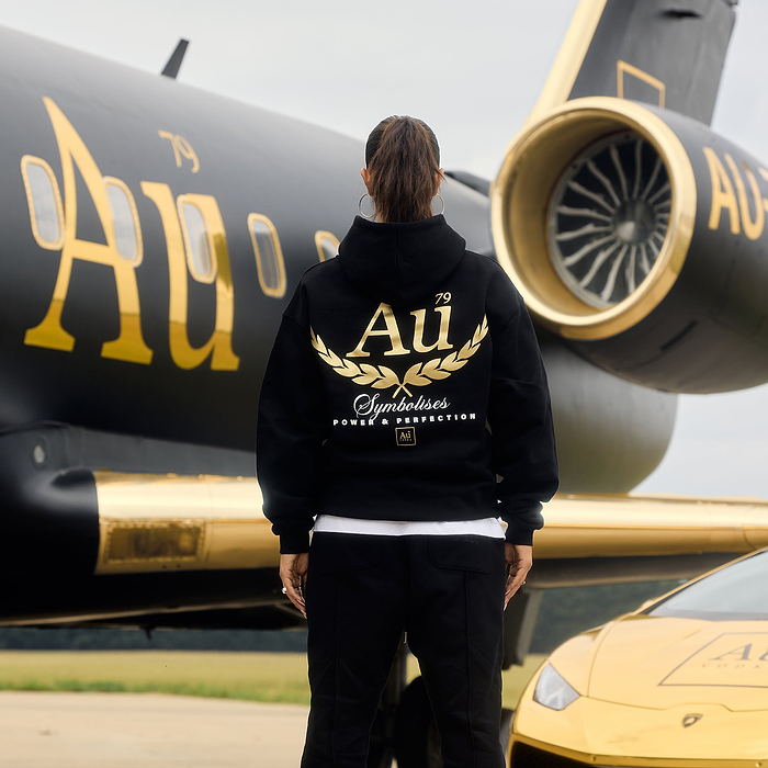 Person wearing a black hoodie with gold 'Au' logo on the back in front of car