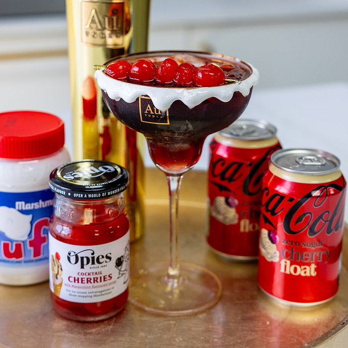 Au Vodka cocktail in a glass rimmed with marshmallow fluff garnished with cherries next to Coca Cola and Opies cherries