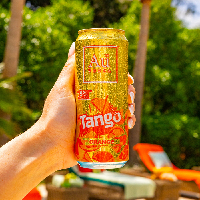 Hand holding a cold Au Vodka Tango Orange can with water droplets, against a blurred green background