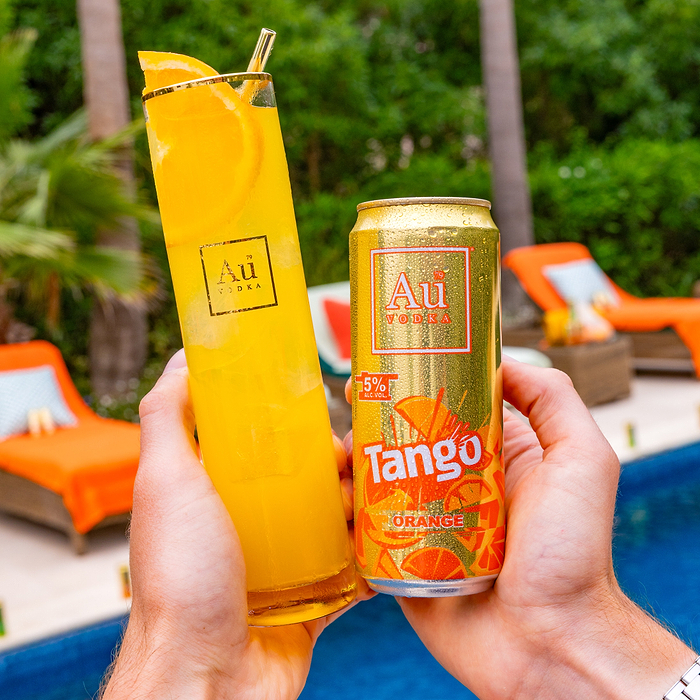 Hands holding an Au Vodka Tango Orange can and a tall glass of orange drink by a pool