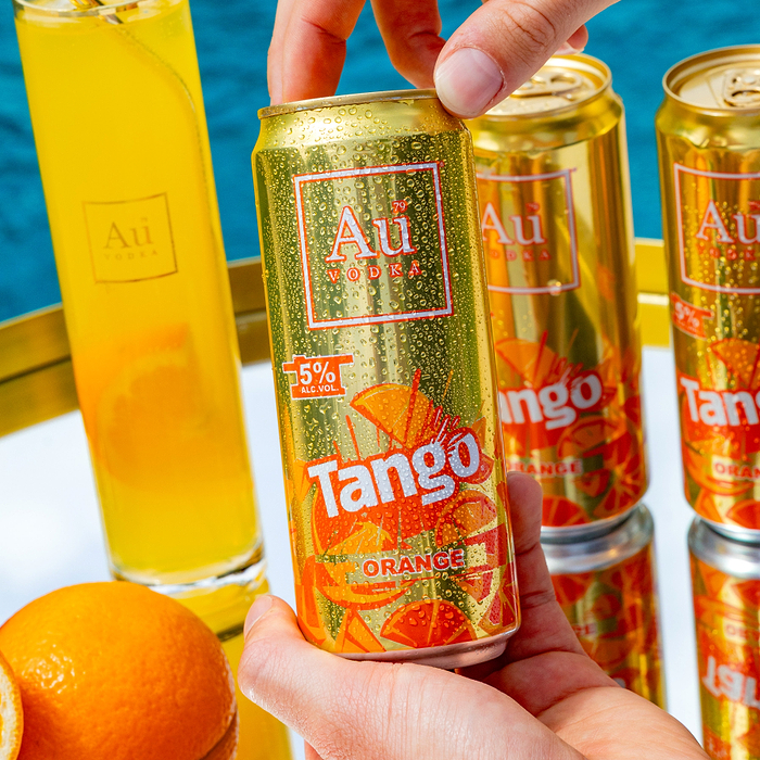 Hand holding a cold Au Vodka Tango Orange can with water droplets, next to other cans and a glass of orange drink