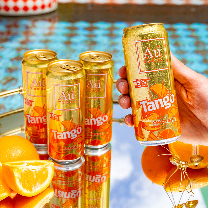 Hand holding a gold Au Vodka Tango Orange can, with three more cans on a mirrored tray with oranges