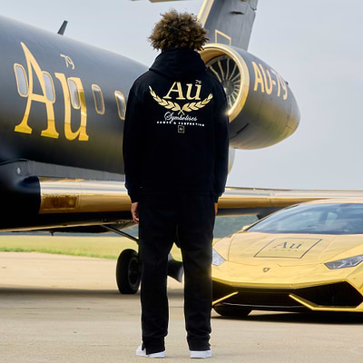 Person wearing a black hoodie with 'Au' logo in front of a black and gold airplane and golf sports car.