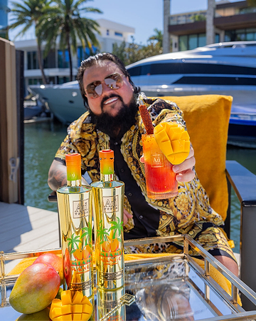 Man in sunglasses holding a mango cocktail, with two Au Vodka Mango bottles and fresh mangoes