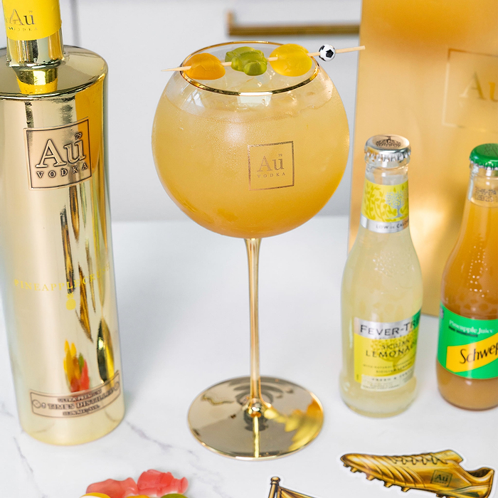Au Vodka Pineapple Crush bottle with a cocktail in a gold rimmed glass garnished with gummy sweets