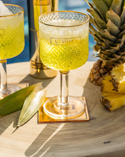 Au Vodka pineapple crush cocktail in a gold-rimmed glass with ice, next to a pineapple and green leaves