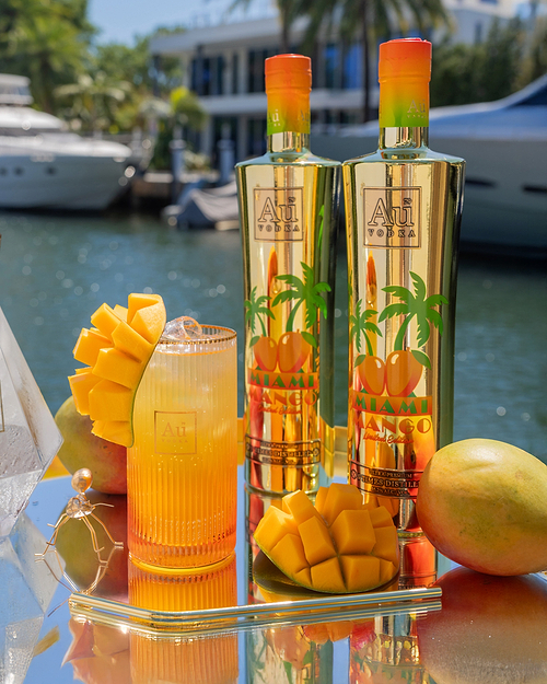 Two Au Vodka Miami Mango bottles, a mango cocktail, and whole and sliced mangoes on a reflective surface