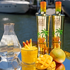 Two Au Vodka Miami Mango bottles, a glass of mango drink, and fresh mangoes on a reflective surface