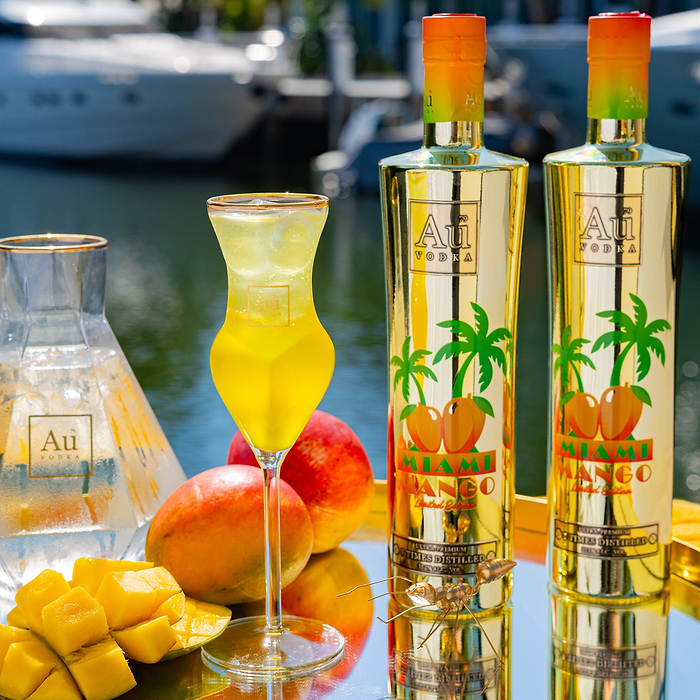 Au Vodka Miami Mango bottles, cocktail, and fresh mango on a reflective surface by the water