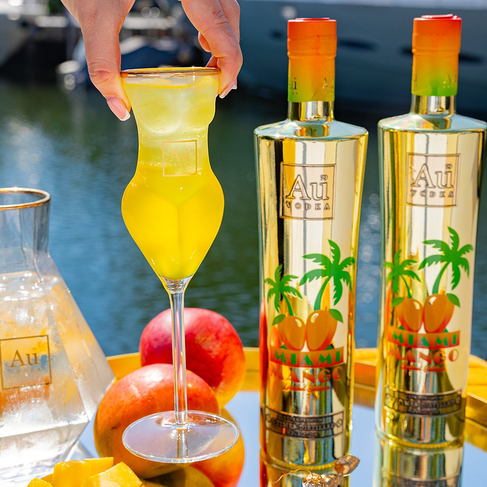 Hand holding a glass of Au Vodka Miami Mango cocktail with two bottles and fresh mangoes