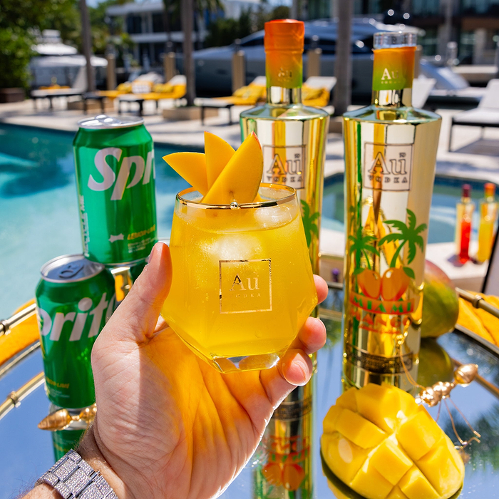 Hand holding a diamond cut glass of Au Vodka Mango Breeze cocktail with mango slices, Sprite cans, and Au Vodka bottles