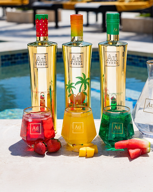 Three Au Vodka bottles and three glasses of cocktails with fruit by a pool
