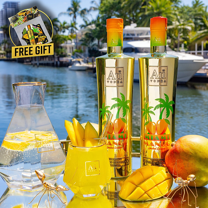 Two Au Vodka Miami Mango bottles, mango slices, and a drink on a reflective surface by a canal