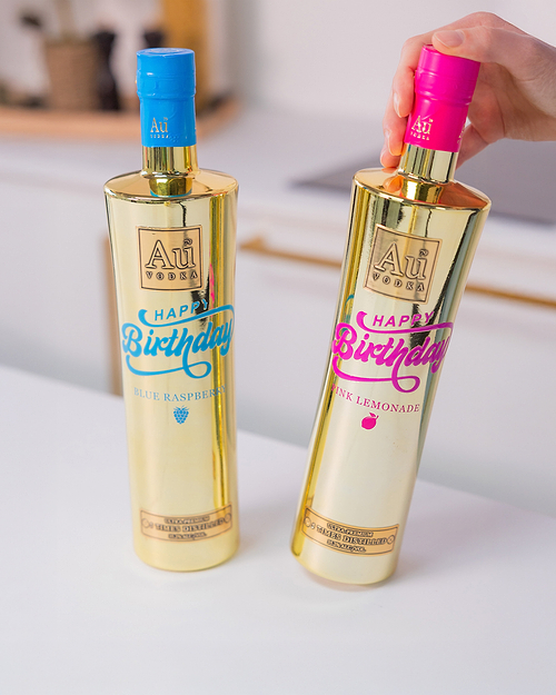 Two Au Vodka Happy Birthday bottles, one blue raspberry and one pink lemonade, on a white surface