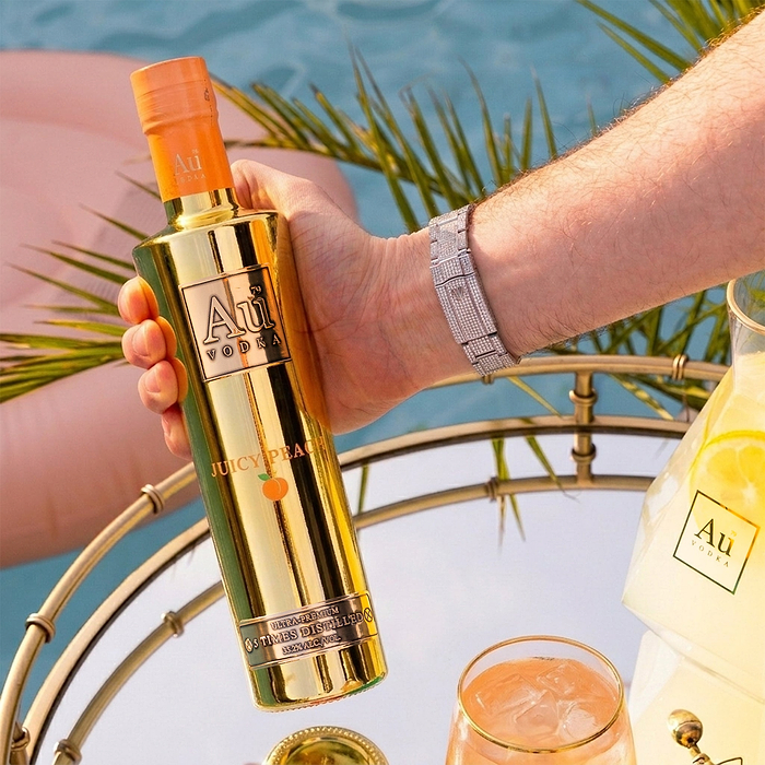 Hand holding a gold Au Vodka Juicy Peach bottle over a mirrored tray with drinks and a pool
