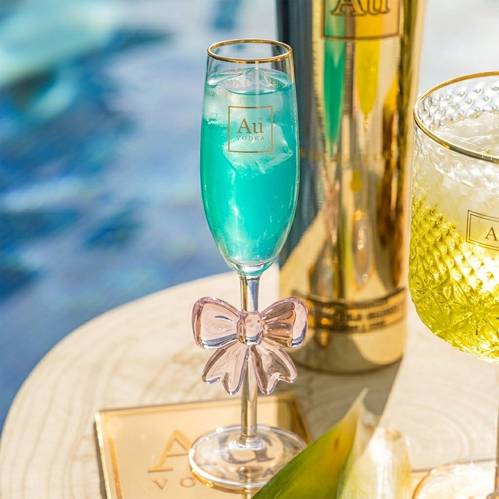 Au Vodka blue cocktail in a champagne flute with a pink bow, next to a gold bottle and yellow drink