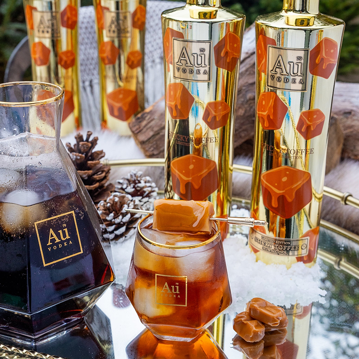Au Vodka bottles with caramel candies and a glass of iced coffee on a reflective surface.