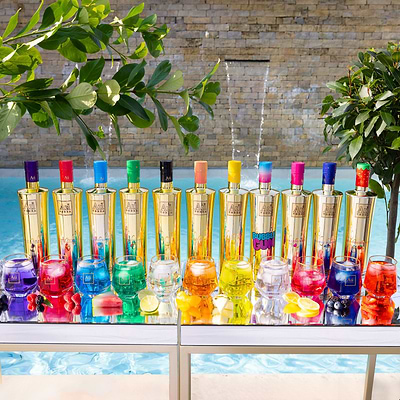 Colorful bottles and glasses on a table by a poolside with plants in the background